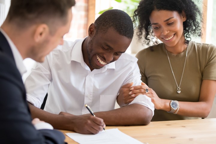 A young man signs a document while a young woman and a man with a suit look on